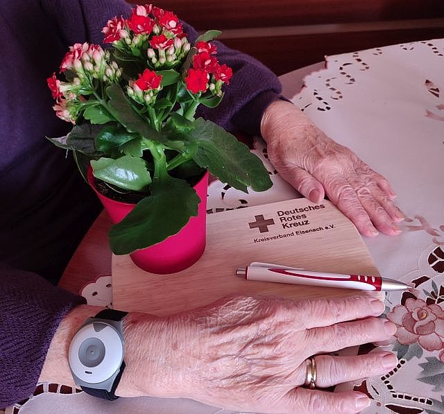 Auf dem Bild sieht man die Hände einer älteren Person, die an einem Tisch sitzt. Die Person trägt an ihrem linken Handgelenk einen Notruf-Knopf. Das ist ein graues, rundes Gerät an einem schwarzen Armband.