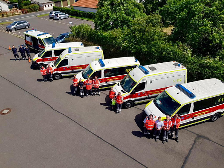 Übersicht von oben: Mitglieder der DRK-Bereitschaft stehen vor einer Reihe von fünf Einsatzfahrzeugen des Katastrophenschutzes und Rettungsdienstes.
