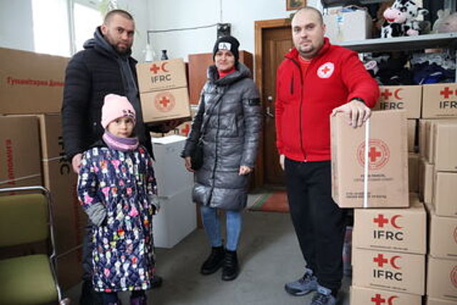 Vier Personen stehen in einem Raum mit vielen braunen Kartons. Ein Mann trägt eine rote Jacke mit einem Kreuz-Symbol. Er hält seine Hand auf einen Stapel Kartons. Auf den Kartons steht „IFRC“ und ein rotes Kreuz. Neben ihm stehen ein weiterer Mann, eine Frau und ein kleines Mädchen in einem bunten Mantel. Sie schauen alle in die Kamera. Im Hintergrund stapeln sich noch mehr Pakete bis an die Wand.