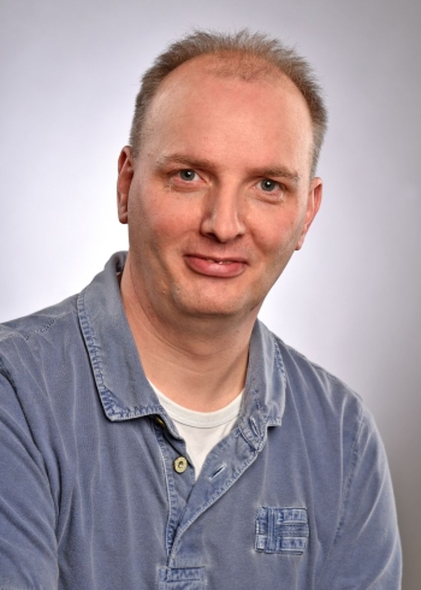 Ein Porträtfoto eines freundlich blickenden Mannes mit kurzen, hellbraunen Haaren. Er trägt ein blaues Poloshirt mit einer dezenten Stickerei auf der Brustseite. Der Hintergrund ist in einem neutralen, hellen Grau gehalten und gleichmäßig ausgeleuchtet. Das Bild wirkt bodenständig, sympathisch und professionell.