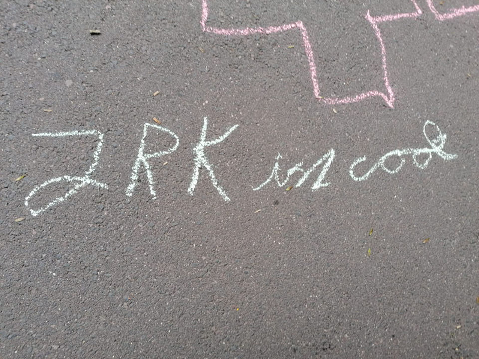 Eine Kreidezeichnung auf grauem Asphalt: In weißer Schrift steht dort der Satz ‚JRK ist cool‘. Darüber ist der Umriss eines großen Kreuzes mit rosa Kreide angedeutet.