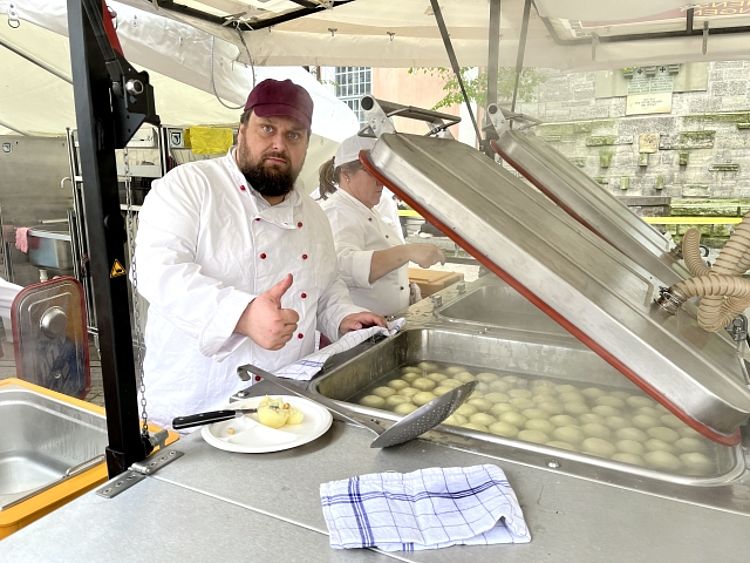 Ein bärtiger Mann in weißer Kochjacke und dunkelroter Mütze steht an einem professionellen, mobilen Kochstand (Feldküche). Er blickt in die Kamera und gibt ein Daumen hoch Geste. Vor ihm befindet sich ein großer Topf mit vielen runden, hellen Kartoffelklößen im kochenden Wasser, sowie ein Teller mit einem aufgeschnittenen Kloß. Im Hintergrund ist eine alte Steinmauer mit Grabtafeln sichtbar und eine weitere Person in Kochkleidung.