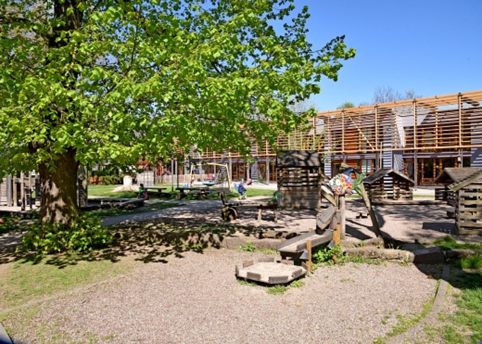 Großzügiges Außengelände des Kindergartens mit altem Baumbestand, Sandkasten und Holzspielhäusern vor dem modernen Hauptgebäude.