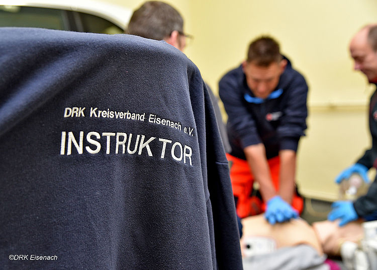 Im Vordergrund ist der Rücken eines dunkelblauen Pullovers zu sehen. Darauf steht in weißer Schrift: „DRK Kreisverband Eisenach e.V. INSTRUKTOR“. Ein Instruktor ist eine Person, die anderen Erste Hilfe beibringt. Im Hintergrund üben zwei Männer an einer Puppe. Einer der Männer drückt mit beiden Händen fest auf den Brustkorb der Puppe. Das ist eine Übung für die Herz-Lungen-Wiederbelebung, um im Notfall Leben zu retten.