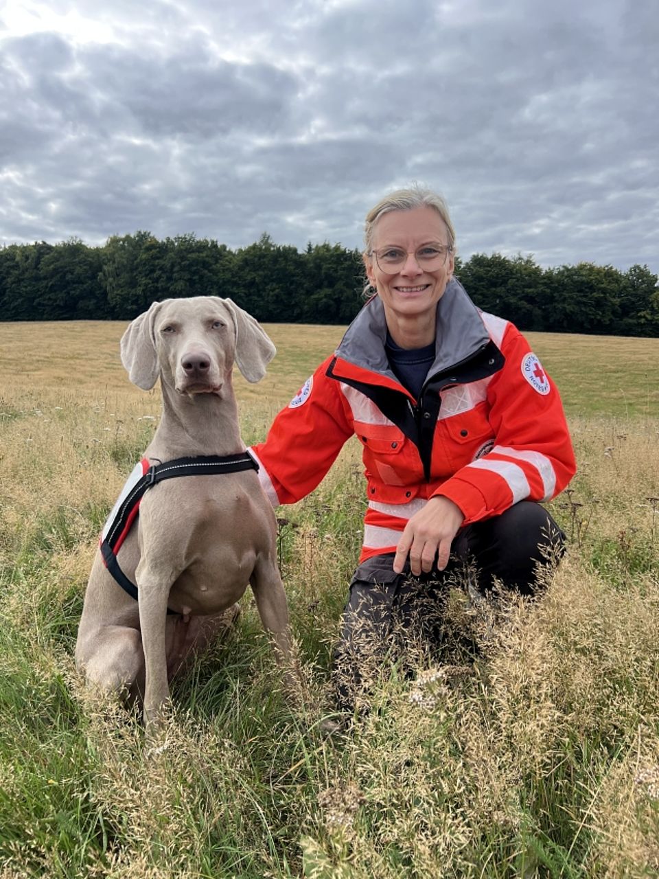 Eine lächelnde, blonde Frau (Rettungskraft) in orange-grauer Einsatzjacke des Deutschen Roten Kreuzes kniet auf einer Wiese neben einem silbrigen Weimaraner Rettungshund. Dieser sitzt ruhig und trägt ein rot-weißes Geschirr mit dem Roten Kreuz. Die Frau legt ihre Hand auf den Rücken des Hundes. Die Szene spielt im Freien vor einem dunklen Waldrand unter einem bewölkten Himmel.