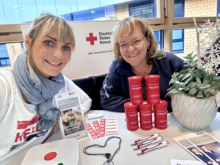 Zwei lächelnde Frauen, Katrin Blaurock und Corina Fitzner, stehen an einem Stand des Deutschen Roten Kreuzes (DRK). Auf dem Tisch vor ihnen liegen Informationsmaterialien, fünf rote Rotkreuzdosen, ein Hausnotrufgerät und Handsender.