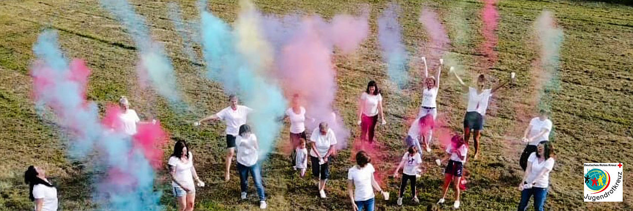 Eine Gruppe von Jugendlichen des Jugendrotkreuzes steht auf einer grünen Wiese und wirft buntes Farbpulver in die Luft. Über den Teilnehmern steigen leuchtende Wolken in Blau, Gelb, Pink und Lila auf. Unten rechts ist das Logo des Jugendrotkreuzes zu sehen.