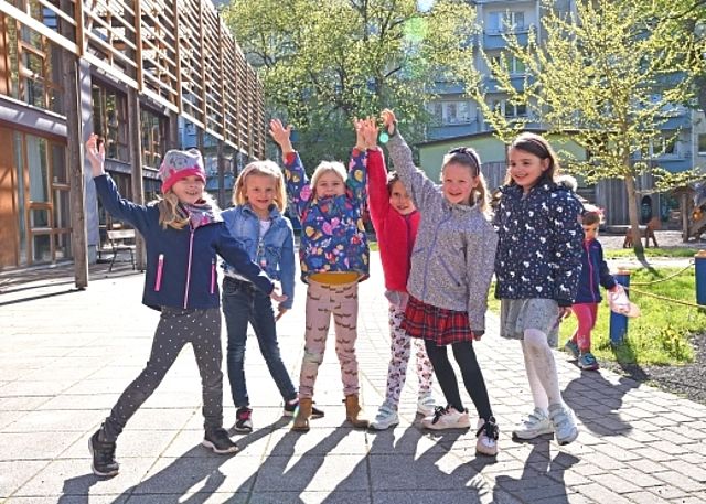 Eine Gruppe fröhlicher Kinder jubelt mit erhobenen Armen im sonnigen Außenbereich des Kindergartens.