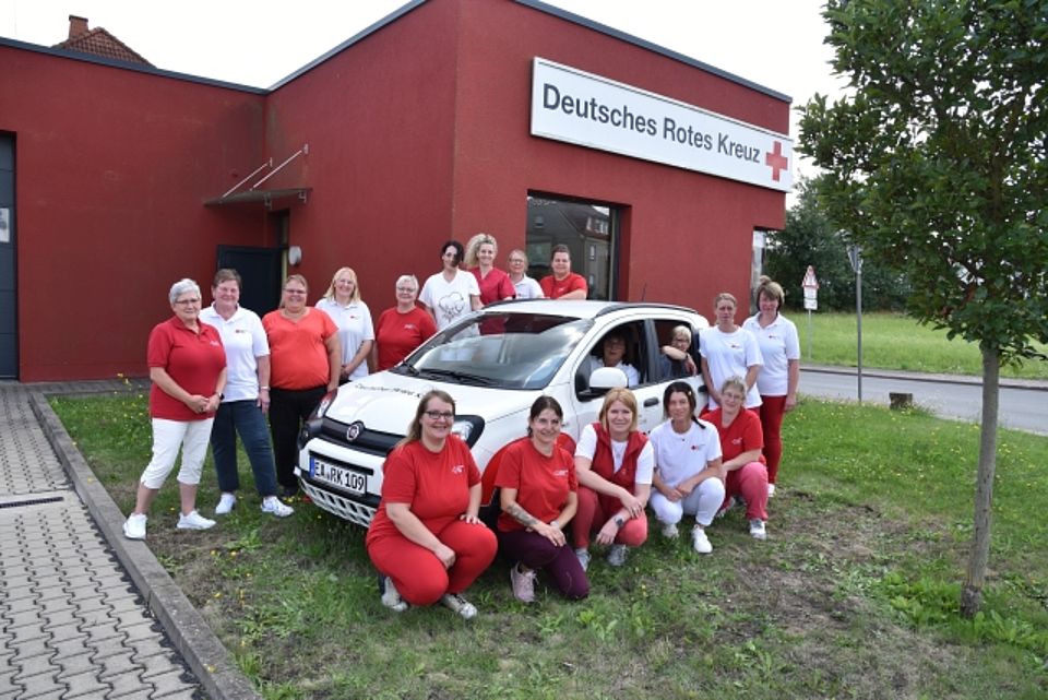 Eine Gruppe von 18 Mitarbeiterinnen des Deutschen Roten Kreuzes (DRK) posiert mit einem weißen DRK-Dienstwagen (Kennzeichen "EA-RK 109") vor einem roten DRK-Gebäude, der Sozialstation Herda. Die Frauen tragen überwiegend rote oder weiße Oberteile sowie Hosen. Alle mit dem DRK-Logo, und sind stehend oder hockend um das Fahrzeug herum gruppiert. Zwei Frauen sitzen im Auto.