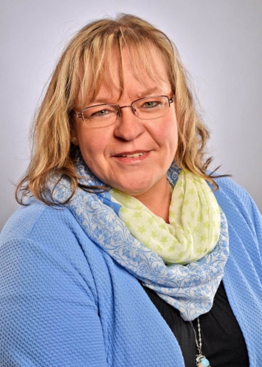 Ein Porträtfoto einer freundlich lächelnden Frau mit längeren, blonden Haaren und einer Brille. Sie trägt eine hellblaue Strickjacke über einem dunklen Oberteil und dazu einen voluminösen, hellgelben Schal. Der Hintergrund ist in einem neutralen, hellen Grau gehalten. Das Bild wirkt herzlich, kompetent und professionell.