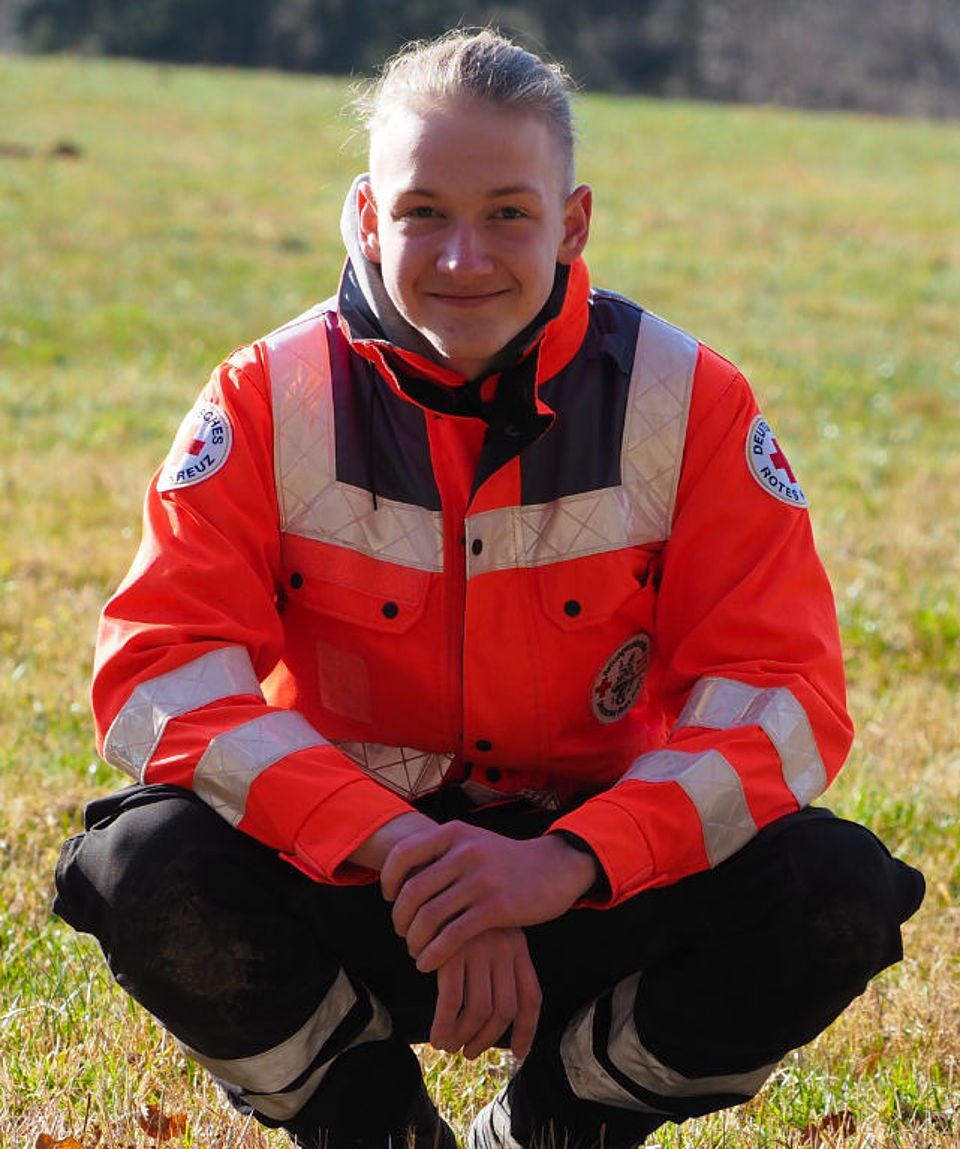 Nahaufnahme eines jungen Mannes (Rettungskraft) mit hellem Haar, das zu einem Zopf zusammengebunden ist. Er hockt lächelnd auf einer Wiese und trägt eine leuchtend orange-graue Einsatzjacke des Deutschen Roten Kreuzes (DRK) mit reflektierenden Streifen. Die Sonne scheint auf ihn, und der Hintergrund ist ein unscharfes, sonniges Feld.