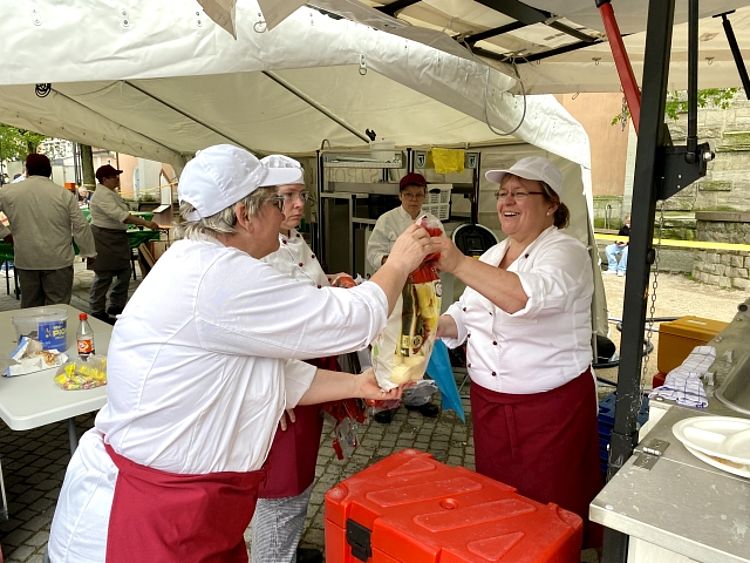 Zwei Frauen in weißen Kochjacken und weißen Kappen, die ehrenamtliche Küchenhelferinnen sind, stehen unter einem weißen Zelt einer Feldküche und reichen sich lachend einen Beutel mit Klößen. Sie tragen dunkelrote Schürzen und arbeiten an einer Essensausgabe im Freien. Im Hintergrund sind weitere Helfer und ein Teil eines historischen Gebäudes zu sehen.