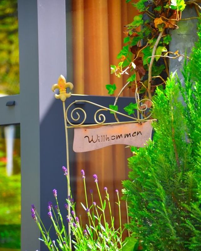 Nahaufnahme eines geschmiedeten Metallstabs, an dem ein geschwungenes Schild mit der Aufschrift „Willkommen“ hängt. Der Stab ist oben mit einer Lilie verziert. Das Schild befindet sich im Außenbereich neben einer gläsernen Türfront. Im Vordergrund blüht violetter Lavendel, und rechts daneben steht ein grüner Nadelstrauch. An der Hauswand rankt Efeu empor. Die Szene wirkt freundlich und einladend.