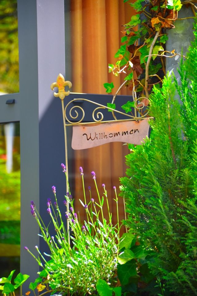 Ein dekoratives, metallenes Willkommensschild in einem Gartenbereich. Das Schild ist an einem Stab befestigt und zeigt den geschwungenen Schriftzug „Willkommen“. Es steht inmitten grüner Pflanzen, darunter ein kleiner Nadelbaum und zarte violette Blüten. Im Hintergrund sind ein Fensterrahmen und ein warmer, orangefarbener Vorhang zu erkennen. Die Szene wirkt einladend und freundlich.