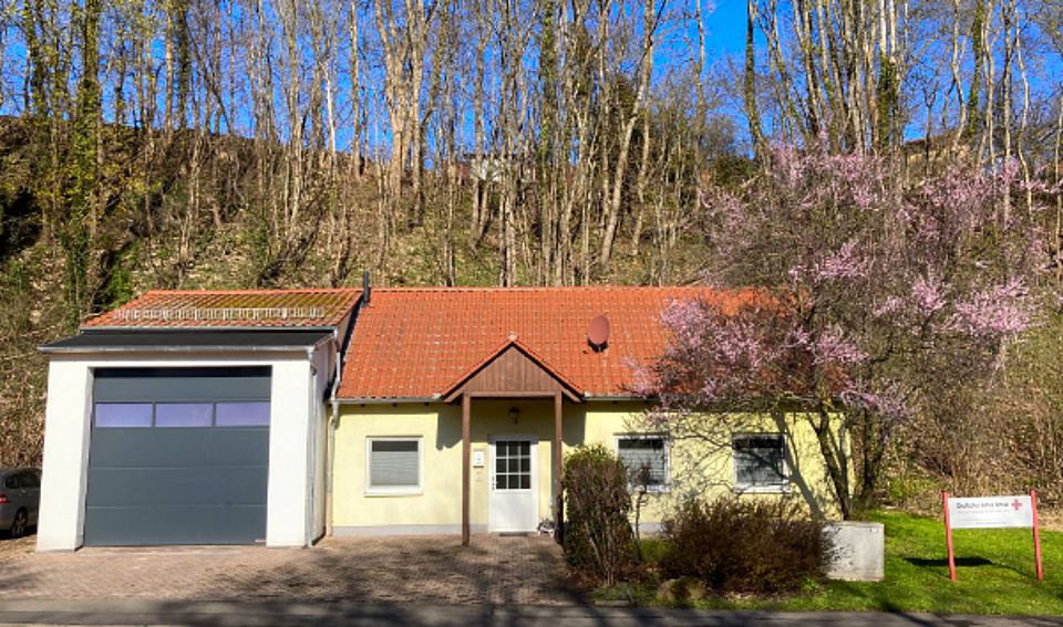 Eine kleine, weiß verputzte Rettungswache mit rotem Ziegeldach und einem Garagenanbau mit dunklem Rolltor. Das Gebäude steht am Fuße eines bewaldeten Hanges. Vor dem Haus blüht ein Baum mit rosafarbenen Blüten.