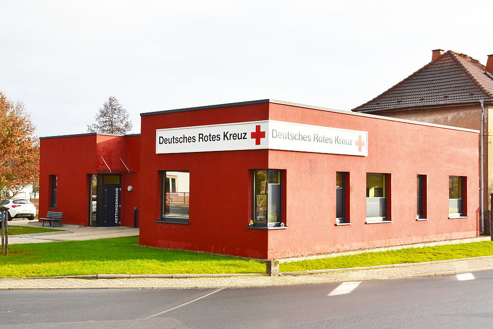 Außenansicht der Rettungswache Herda des Deutschen Roten Kreuzes (DRK). Das moderne, quaderförmige Gebäude ist in einem kräftigen Rotton gehalten und trägt über den Fenstern einen weißen Schriftzug mit dem Namen und dem Logo des Deutschen Roten Kreuzes.