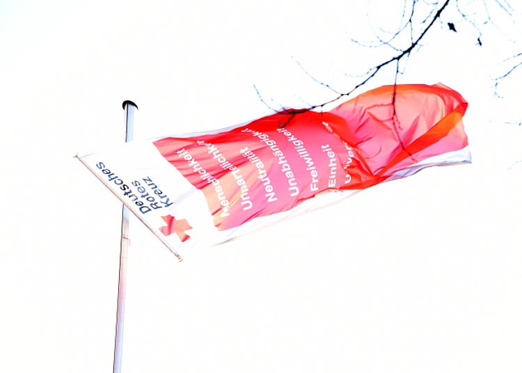 Eine weiße, längliche Fahne des Deutschen Roten Kreuzes (DRK), die im Wind vor einem hellen Himmel weht. Auf dem Teil, der am Fahnenmast hängt, ist das Logo und der Schriftzug "Deutsches Rotes Kreuz" zu sehen. Im roten Teil der Fahne sind die sieben Grundsätze des DRK, Menschlichkeit, Unparteilichkeit, Neutralität, Unabhängigkeit, Freiwilligkeit, Einheit und Universalität in weißer Schrift erkennbar.