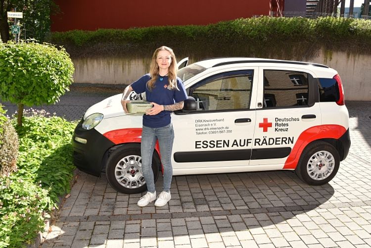 Eine engagierte junge Helferin des Deutschen Roten Kreuzes strahlt freundlich und hält sorgfältig eine warme Essensbox in den Händen, bereit für die tägliche Auslieferung. Sie steht selbstbewusst vor dem DRK-Fahrzeug, das deutlich mit 'ESSEN AUF RÄDERN' beschriftet ist und symbolisiert damit die verlässliche und herzliche Unterstützung, die älteren und bedürftigen Menschen nach Hause gebracht wird.
