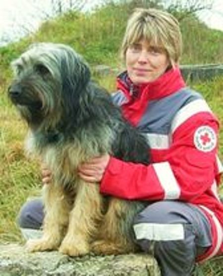 Eine blonde Rettungskraft in einer rot-grauen Einsatzjacke mit dem Emblem des Deutschen Roten Kreuzes hockt neben einem großen, langhaarigen Schäferhund oder Briard-ähnlichen Rettungshund. Sie hält den Hund umarmt fest und blickt direkt in die Kamera. Der Hund blickt seitlich nach links. Der Hintergrund ist eine unscharfe, grüne Hügellandschaft.