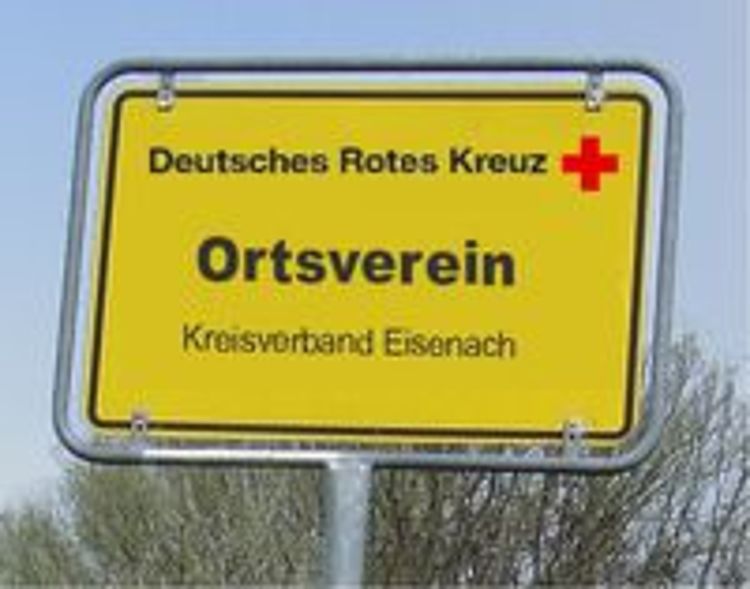 Ein quadratisches, gelbes Schild an einem Pfosten imitiert das Design eines deutschen Ortseingangsschildes. Darauf steht in schwarzen Buchstaben: "Deutsches Rotes Kreuz" mit einem roten Kreuz-Symbol. Darunter steht in großer Schrift "Ortsverein", und ganz unten "Kreisverband Eisenach". Das Schild steht im Freien unter einem hellblauen Himmel.