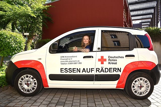 Ein weißes Auto steht auf der Straße. Auf der Tür steht groß: ESSEN AUF RÄDERN. Daneben sieht man das Logo vom Deutschen Roten Kreuz. Eine Frau sitzt am Steuer und lächelt. Sie hält eine Box mit Essen in der Hand. Im Hintergrund sind grüne Bäume und ein rotes Haus.