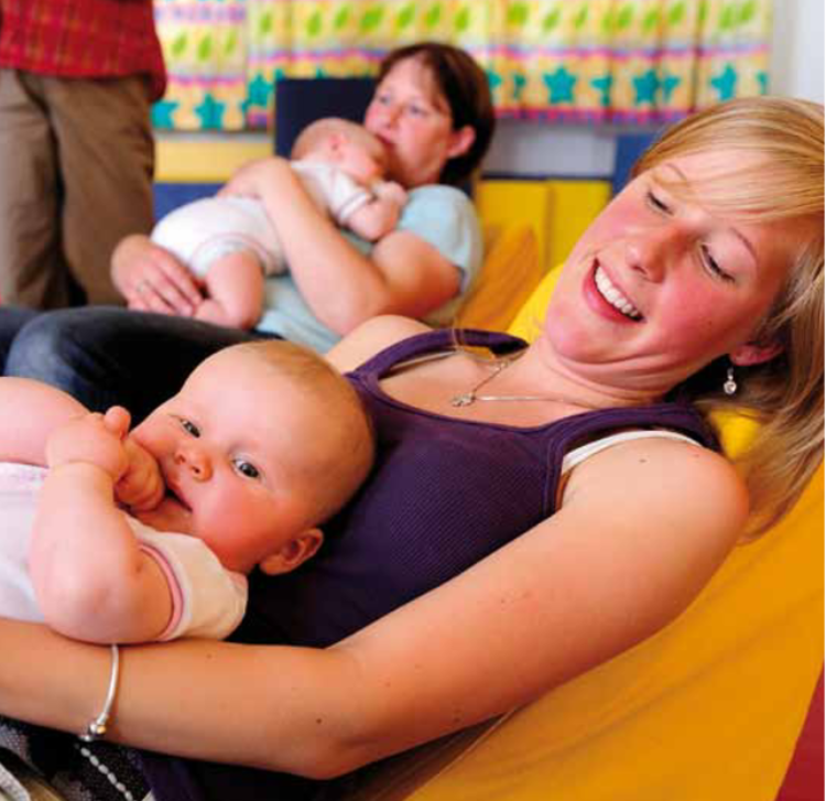 Eine helle, freundliche Szene in einem Gruppenraum, in dem zwei Frauen mit ihren Babys zu sehen sind. Im Vordergrund sitzt eine lächelnde Frau in einem gelben Sitzsack und hält ein waches Baby auf dem Schoß, das neugierig in die Kamera blickt. Im Hintergrund sitzt eine weitere Frau auf einem blauen Polster und wiegt ein schlafendes Baby im Arm. Die Atmosphäre wirkt entspannt und gemeinschaftlich; an der Wand hängt eine bunte Girlande mit Sternenmotiven.