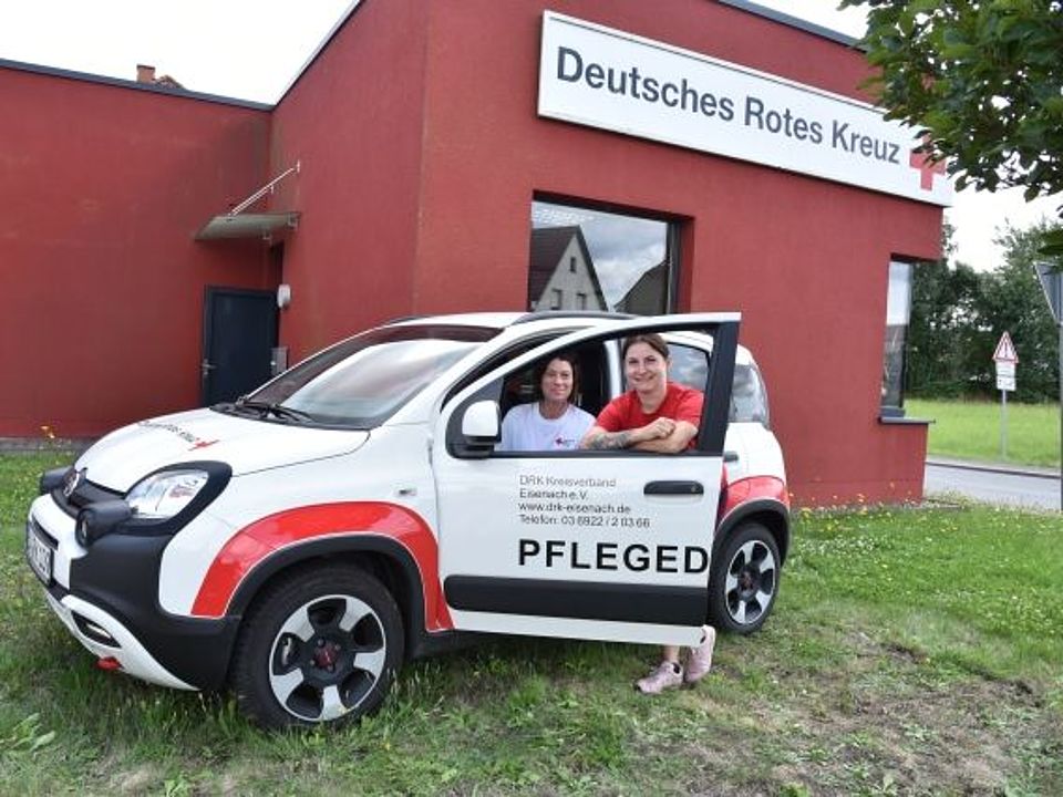 Weißes Dienstfahrzeug des Deutschen Roten Kreuzes (DRK) mit der Aufschrift Pflegedienst in Eisenach. Zwei lächelnde Mitarbeiterinnen – eine am Steuer, die andere an der offenen Beifahrertür – posieren vor einem roten DRK-Gebäude, der Sozialstation in Herda.
