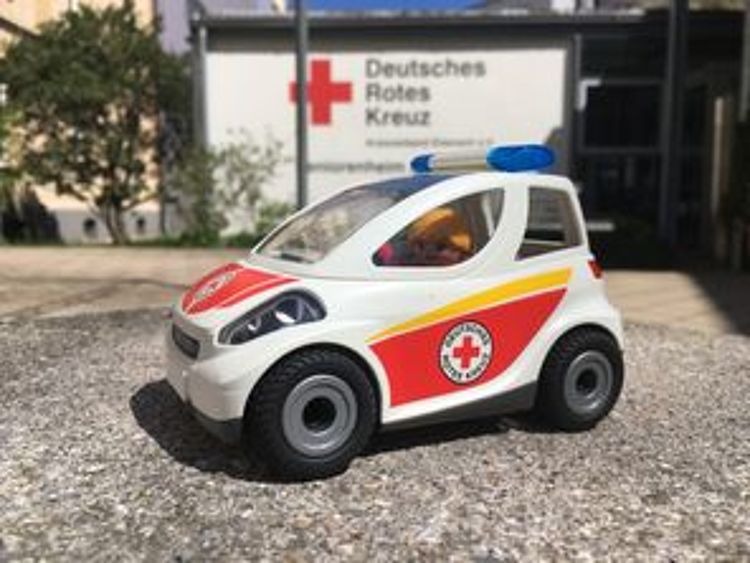 Ein kleines Spielzeug-Auto von Playmobil steht draußen auf einem Stein-Boden. Das Auto ist weiß und hat rote Flächen mit dem Logo vom Deutschen Roten Kreuz. Auf dem Dach ist ein blaues Licht. Eine kleine Figur mit blonden Haaren sitzt am Steuer. Im Hintergrund sieht man unscharf ein Gebäude vom Deutschen Roten Kreuz.