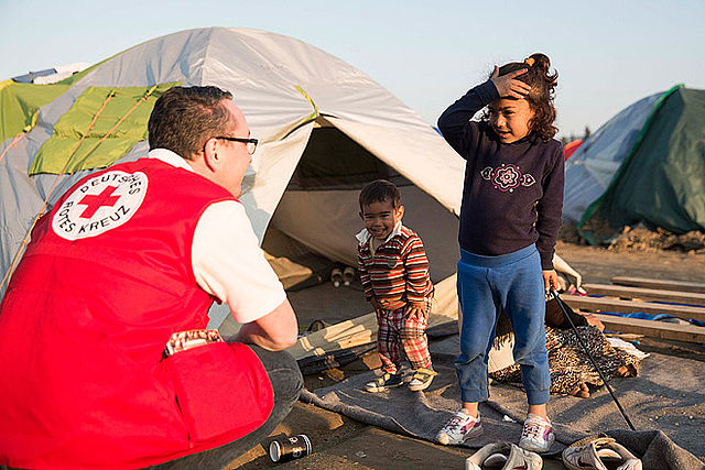 Ein Helfer vom Deutschen Roten Kreuz trägt eine rote Weste. Er hockt vor einem Zelt und spricht mit zwei Kindern. Ein kleiner Junge lacht im Zelteingang. Ein Mädchen steht daneben und hält sich die Hand an den Kopf. Im Hintergrund stehen weitere Zelte auf dem Boden.