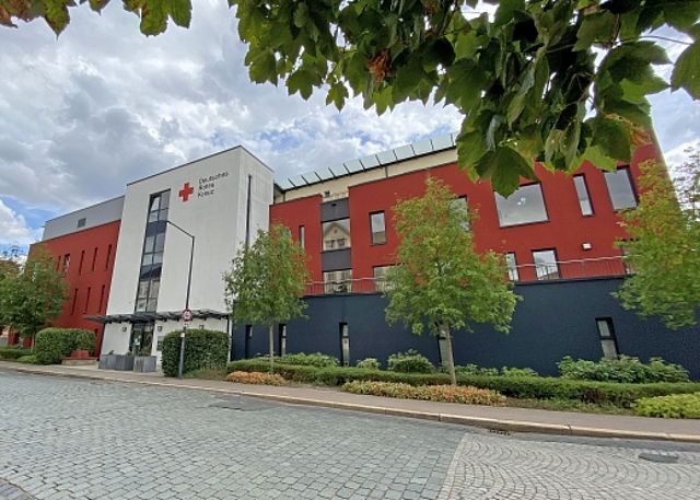 Vorderansicht Haus Fischerstadt. Das Bild zeigt ein großes, modernes Haus an einer Straße mit Pflaster-Steinen. Das Haus hat drei Farben: Weiß, Rot und Dunkel-Blau. Auf dem weißen Teil oben steht „Deutsches Rotes Kreuz“ mit einem roten Kreuz daneben. Vor dem Haus wachsen grüne Hecken und kleine Bäume. Der Himmel ist hell mit ein paar Wolken.