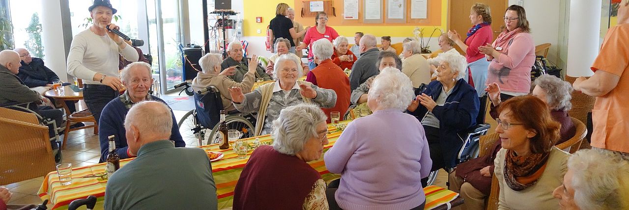 Ein Fest im Seniorenwohnheim Justusstift. Viele Seniorinnen und Senioren sitzen gemeinsam an Tischen. Ein Alleinunterhalter mit weißem Shirt und Hut singt und unterhält die Senioren. Pflegekräfte und Gäste stehen im Raum. Die Stimmung ist fröhlich.