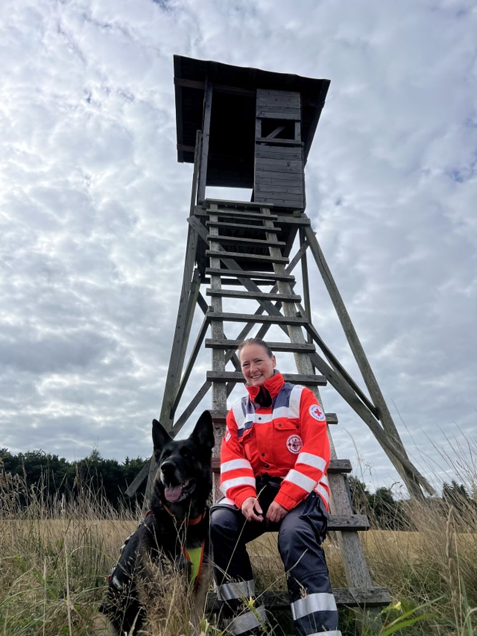 Eine lächelnde Frau (Rettungskraft) in orange-grauer Einsatzkleidung des Deutschen Roten Kreuzes sitzt auf einer Sprosse einer hohen, dunklen Holzleiter, die zu einem Hochsitz führt. Neben ihr sitzt ein großer, schwarzer Schäferhund Rettungshund mit abstehenden Ohren und geöffnetem Maul. Die Szene ist im Freien auf einer trockenen Wiese vor einem Waldrand und unter einem dramatisch bewölkten Himmel aufgenommen.