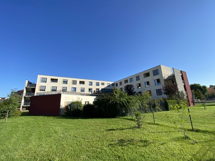 Das Bild zeigt ein sehr großes, helles Haus mit vielen Fenstern. Das Haus hat die Form von einem L. Vor dem Haus ist eine sehr große, grüne Wiese. Es gibt einen Zaun und einige grüne Büsche und kleine Bäume. Der Himmel ist ganz blau und es gibt keine Wolken. Die Sonne scheint hell auf das Gebäude und das Gras.