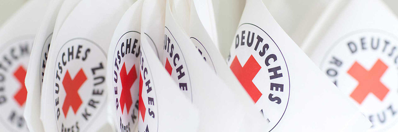 Eine Nahaufnahme mehrerer kleiner, weißer Wimpel oder Flaggen, die dicht nebeneinander hängen. Auf jeder Flagge ist das Logo des Deutschen Roten Kreuzes zu sehen: ein rotes Kreuz in einem Kreis, umrandet mit dem schwarzen Schriftzug „DEUTSCHES ROTES KREUZ“. Der Fokus liegt auf den vorderen Logos, während der Hintergrund leicht unscharf wirkt.
