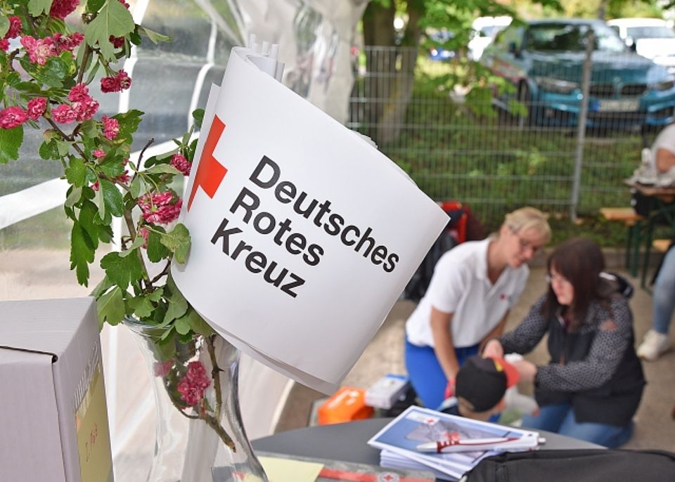Nahaufnahme eines weißen Wink Elements des Deutschen Roten Kreuzes (DRK) mit dem roten Kreuz-Symbol und dem Schriftzug "Deutsches Rotes Kreuz", befestigt an einem Strauß mit rosafarbenen Blüten. Im unscharfen Hintergrund kniet eine Ausbilderin in Erster Hilfe (in weißem DRK-Poloshirt) neben einer Besucherin (Mutter) und ihrem Sohn am Stand eines Informationstages. Die Ausbilderin demonstriert und übt mit dem Jungen eine Erste-Hilfe-Maßnahme.