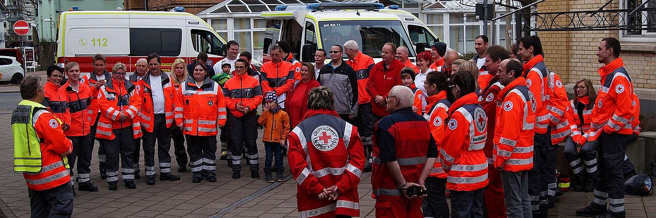 Eine große Gruppe von DRK-Helfern, bestehend aus etwa 20 bis 25 Personen, steht eng zusammen auf einem gepflasterten Platz. Die meisten tragen leuchtend orange-rote Einsatzjacken mit reflektierenden Streifen und DRK-Emblemen. Einige wenige tragen andere DRK-Kleidung oder haben Warnwesten an. Im Hintergrund sind zwei Krankenwagen und Stadtgebäude zu sehen. Die Gruppe scheint sich für eine Besprechung oder ein Gruppenfoto versammelt zu haben.