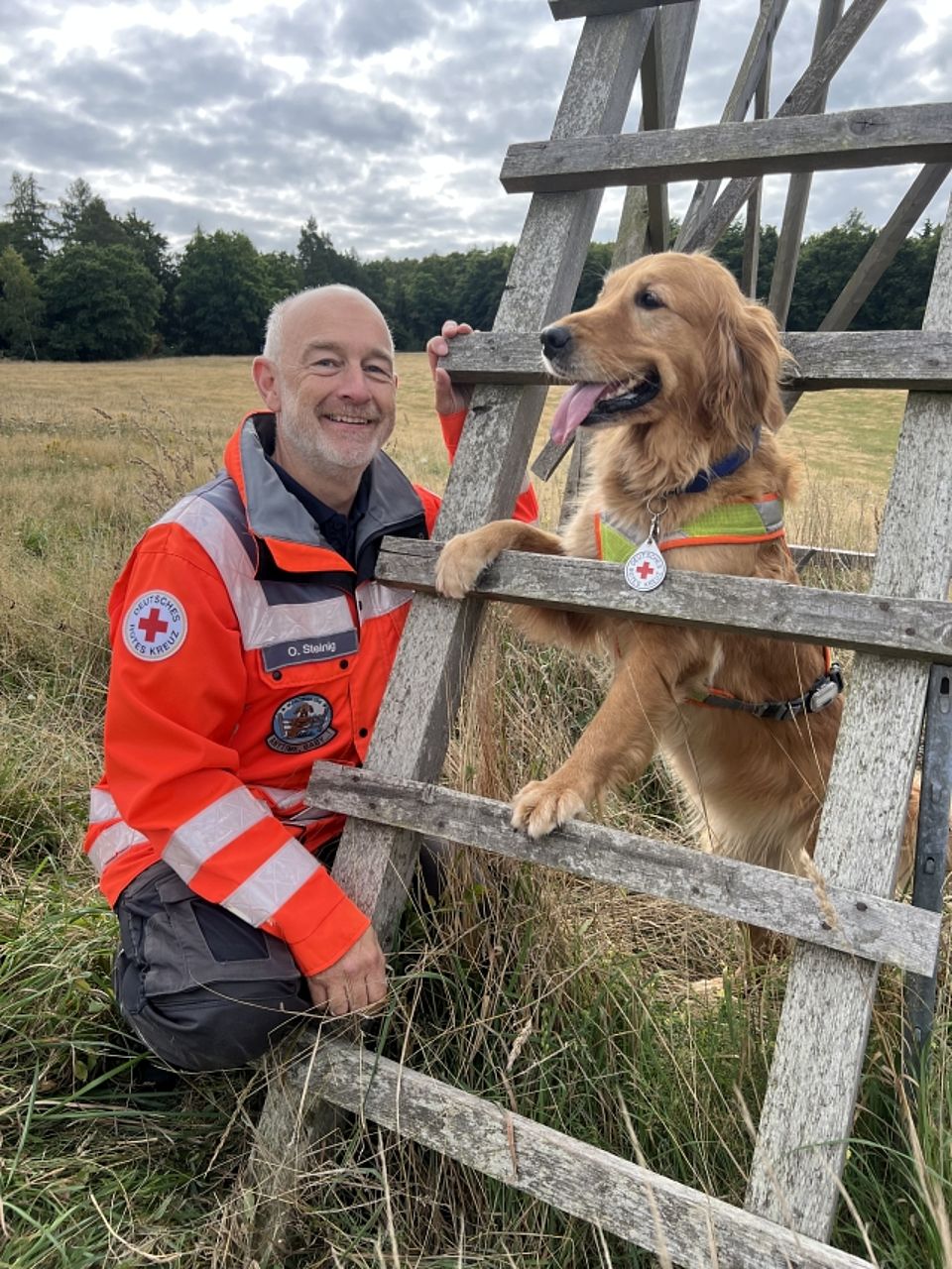Ein freundlich lächelnder, kurzhaariger Mann (eine Rettungskraft mit dem Namensschild "O. Steinig") kniet in orangefarbener Einsatzjacke des Deutschen Roten Kreuzes neben einem hellbraunen Golden Retriever Rettungshund. Der Hund steht mit den Vorderpfoten auf einer rustikalen Holzleiter oder einem Zaun und trägt eine reflektierende Weste mit einer Plakette des Roten Kreuzes. Die Szene spielt draußen auf einer Wiese mit Wald im Hintergrund.