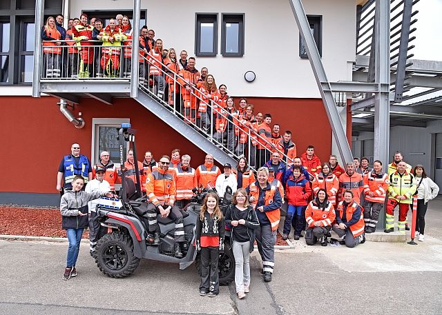 Teamfoto des DRK Eisenach: Zahlreiche ehrenamtliche Helfer in orangefarbener Einsatzkleidung posieren auf und vor einer Außentreppe der Rettungswache. Im Vordergrund steht ein graues Rettungs-Quad.