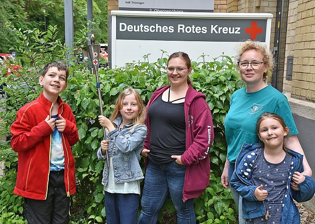 Zwei Frauen und drei Kinder stehen gemeinsam vor einer grünen Hecke und lächeln in die Kamera. Im Hintergrund ist ein großes weißes Schild mit der Aufschrift „Deutsches Rotes Kreuz“ und dem roten Kreuz-Logo zu sehen. Die Gruppe ist sommerlich gekleidet, unter anderem in einer roten Jacke und einem türkisfarbenen T-Shirt. Das Foto vermittelt eine freundliche, familiäre Atmosphäre vor einem DRK-Gebäude.