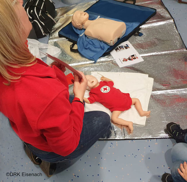Eine Frau in einer roten Jacke sitzt auf dem Boden und hält ein rotes Kopfschnittmodell in der Hand. Vor ihr liegen zwei Übungspuppen auf einer glänzenden Matte: eine größere Puppe „Junior“ und eine kleine Puppe „Baby“ in einem roten Anzug. Die Frau erklärt mit dem Modell, wie man die Atemwege frei macht, um bei Kindern und Babys im Notfall richtig zu helfen.