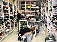 Zwei Frauen arbeiten in einem hellen Raum mit vielen weißen Regalen. Die Regale sind bis oben hin voll mit ordentlich gestapelter Kleidung. In der Mitte steht ein großer Arbeitstisch. Dort sortieren die Frauen weitere Kleidungsstücke. Überall in den Regalen und auf dem Boden stehen Kisten und Körbe mit Wäsche.
