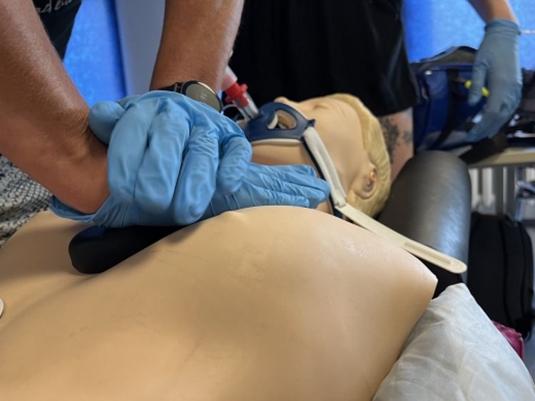 Eine Nahaufnahme zeigt zwei Hände in blauen Schutzhandschuhen, die eine Herzdruckmassage bei einer Übungspuppe durchführen. Die Hände sind fest in der Mitte des Brustkorbs übereinandergelegt. Die Puppe trägt eine dunkle Halskrause zur Stabilisierung. Im Hintergrund ist eine weitere Person in dunkler Kleidung zu sehen, die ebenfalls blaue Handschuhe trägt.