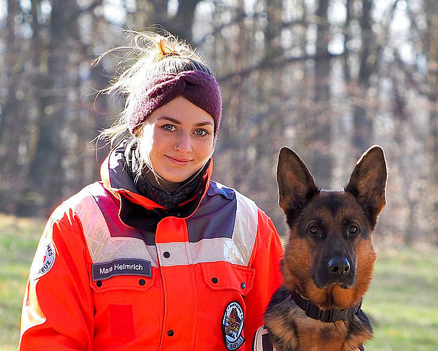 Ein Porträt einer jungen Rettungshundeführerin des Deutschen Roten Kreuzes und ihres Deutschen Schäferhundes vor einem herbstlichen Waldhintergrund. Die Frau trägt eine leuchtend orangefarbene Einsatzjacke mit Reflektoren, ein Namensschild und ein dunkles Stirnband. Sie blickt lächelnd in die Kamera. Rechts neben ihr sitzt der Hund aufmerksam und schaut ebenfalls direkt in die Kamera. Die Szene wirkt professionell und zeigt die enge Bindung zwischen Hundeführerin und Suchhund im DRK-Kreisverband Eisenach e.V.