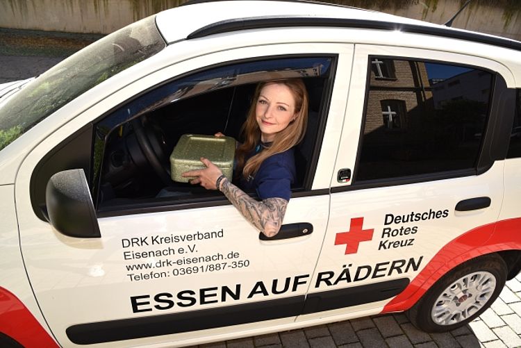 Dieses Bild zeigt eine freundlich lächelnde Frau mit Tätowierungen am Arm, die aus dem Fenster eines weißen Lieferwagens des Deutschen Roten Kreuzes (DRK) heraus eine Essensbox hält. Der Schriftzug auf dem Fahrzeug identifiziert es als Dienstfahrzeug des DRK Kreisverband Eisenach e.V. für den Dienst "ESSEN AUF RÄDERN" (Menüservice). Die Aufschriften beinhalten auch die Kontaktdaten: •	Website: drk-eisenach.de •	Telefon: 03691/887-350 Die Frau scheint eine Mitarbeiterin oder ehrenamtliche Helferin des Menüservices zu sein, die gerade eine Mahlzeit ausliefert. Die Gesamtstimmung des Bildes ist hilfsbereit, freundlich und engagiert. Der Service "Essen auf Rädern" des DRK Kreisverband Eisenach liefert täglich oder wöchentlich warme Mittagsmenüs oder tiefgekühlte Mahlzeiten an Menschen, die sich nicht selbst versorgen können oder möchten, insbesondere an Senioren.