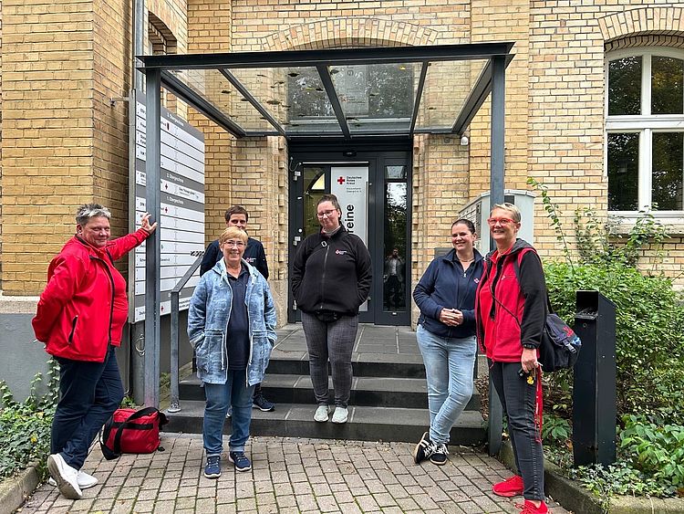 Sechs Erwachsene, einige in roten Jacken, stehen vor dem verglasten Eingang eines Backsteingebäudes. Im Hintergrund ist ein Schild mit dem Logo des Roten Kreuzes zu erkennen.