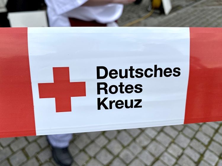 Nahaufnahme eines horizontal gespannten rot-weißen Absperrbandes des Deutschen Roten Kreuzes (DRK). Auf dem Band ist links das rote Kreuz auf weißem Grund abgebildet, und daneben steht in schwarzen Buchstaben der Text: "Deutsches Rotes Kreuz". Im unscharfen Hintergrund ist ein Teil eines Kopfsteinpflasters und die Beine einer Person in weißer Kleidung zu sehen.
