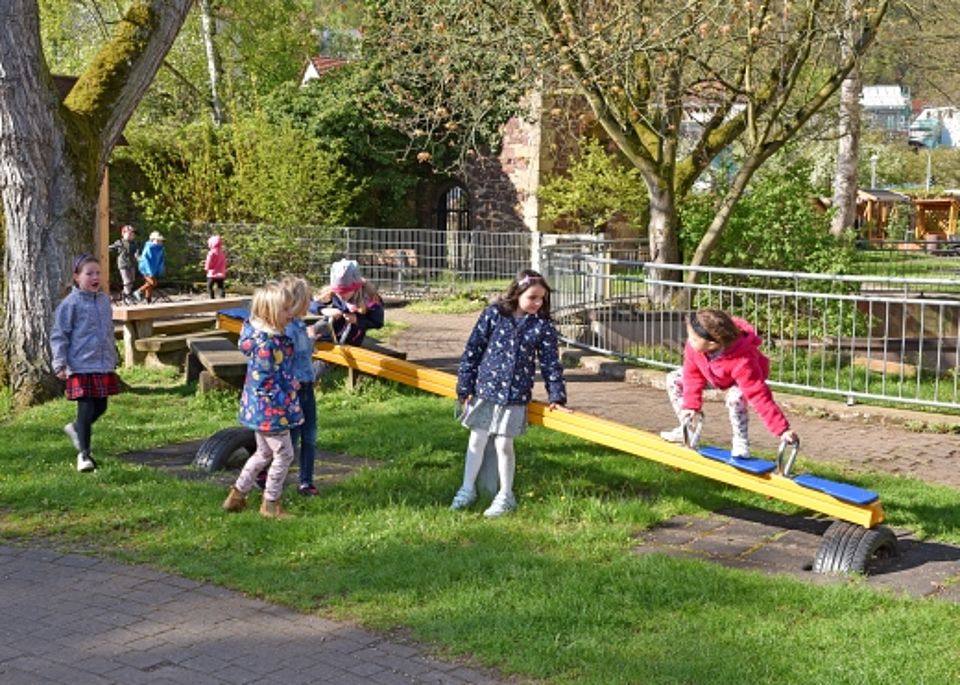 Kinder spielen gemeinsam auf einer gelben Wippe im grünen Außengelände des Kindergartens.