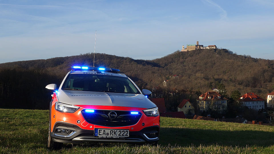 Ein weises Auto vom Roten Kreuz steht auf einer Wiese. Es ist ein Notarzt-Einsatz-Fahrzeug. Auf dem Dach und vorne am Auto leuchten helle, blaue Lichter. Im Hintergrund sieht man einen Berg mit vielen Bäumen. Ganz oben auf dem Berg steht eine große, alte Burg. Das ist die Wartburg in Eisenach.