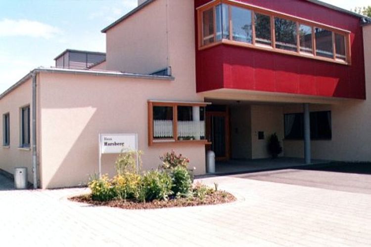 Das moderne Gebäude des "Haus Harsberg", einer DRK-Einrichtung für Menschen mit seelischer und geistiger Behinderung in Mihla. Die Fassade ist hellbeige. Der Eingangsbereich wird von einem auffälligen, tiefroten Balkon-Vorbau überdacht. Auf dem gepflasterten Vorplatz steht ein Schild mit dem Namen der Einrichtung, umgeben von einer kleinen Blumeninsel.
