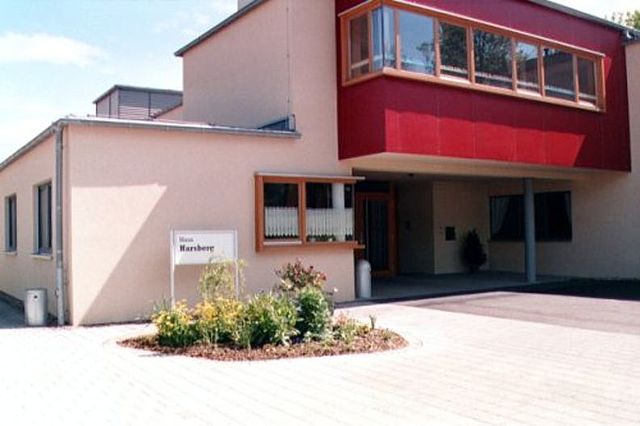 Das Bild zeigt den Eingangsbereich vom Haus Harsberg. Das Gebäude hat helle Wände und einen modernen Anbau in dunkler roter Farbe mit vielen Fenstern. Vor dem Eingang ist ein gepflasterter Platz. Ein kleines Blumenbeet mit bunten Pflanzen verschönert den Bereich. Links neben den Blumen steht ein weißes Schild, auf dem „Haus Harsberg“ steht.